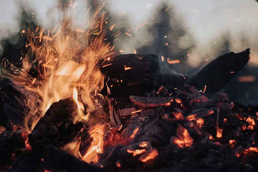 Bild von einem heruntergebrannten Feuer, in dem noch einzelne Holzstücke ersichtlich sind und noch einige wenige Flammen lodern.