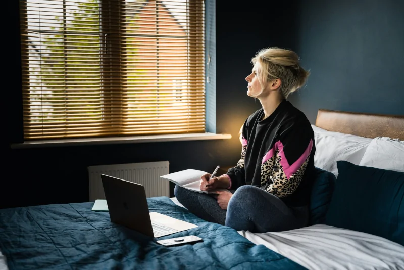 Frau sitzt mit ihrem Laptop und Handy im Bett und arbeitet als Sinnbild für Perfektionismus