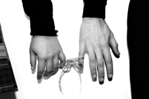 Blick auf die Handrücken von zwei Menschen, die nebeneinander positioniert sind. Die kleinen Finger der beiden Hände sind mit einem Faden, der zu einer Masche geformt ist, verbunden.